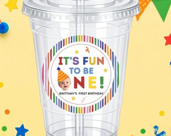 May include: Clear plastic cup with a lid and straw. A colorful sticker on the cup reads "IT'S FUN TO BE ONE!" with a picture of a baby wearing a party hat. The text "BRITTANY'S FIRST BIRTHDAY" is also on the sticker. The background is yellow with party decorations.