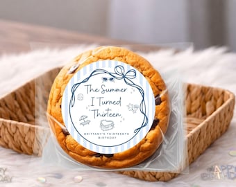 May include: A large chocolate chip cookie in a clear cellophane bag. The cookie has a round sticker with blue stripes and the text "The Summer I Turned Thirteen". The cookie is in a woven basket on a white surface.