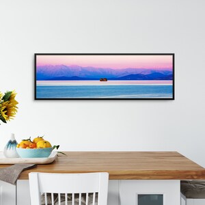 May include: A panoramic photograph of a mountain range with a small island in the middle of a lake. The sky is a vibrant pink and purple, and the water is a deep blue. The photograph is framed in a black frame.