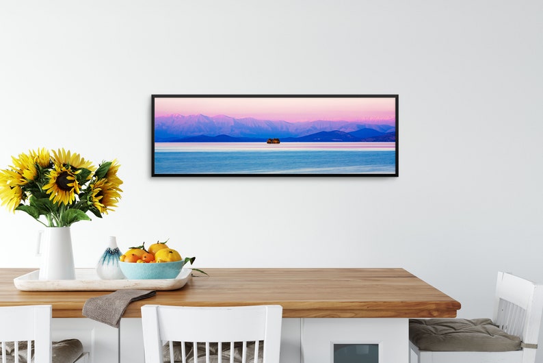 May include: A panoramic photograph of a mountain range with a small island in the middle of a lake. The sky is a vibrant pink and purple, and the water is a deep blue. The photograph is framed in a black frame.