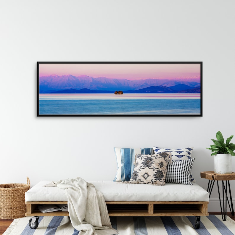May include: A panoramic photograph of a mountain range in the distance, with a small island in the middle of a body of water. The sky is a vibrant pink and purple, and the water is a deep blue.