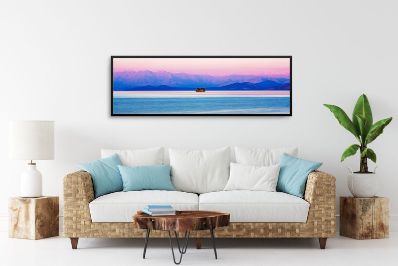 May include: A panoramic photograph of a mountain range with a small island in the middle of a lake. The sky is a vibrant pink and purple, and the water is a deep blue. The image is framed in a black frame.