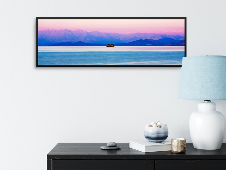 May include: A panoramic photograph of a mountain range with a pink and purple sky at sunset. A small island with trees is in the middle of a calm lake in the foreground.