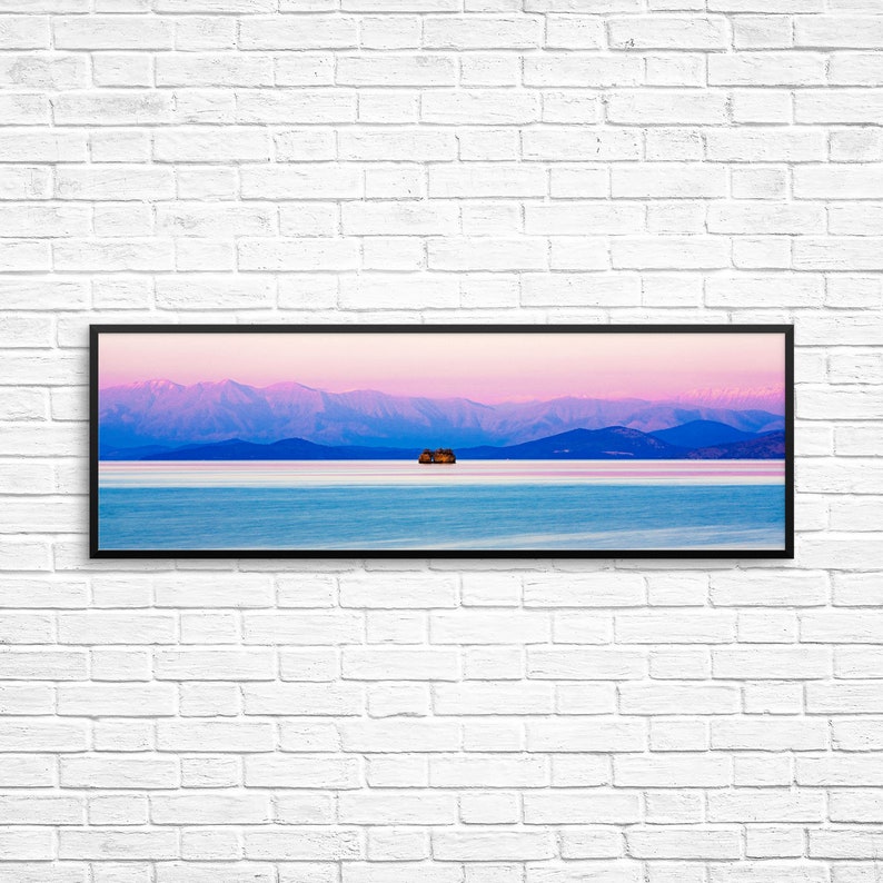 May include: A panoramic view of a small island with trees in a body of water with a mountain range in the background. The sky is a gradient of pink and blue.
