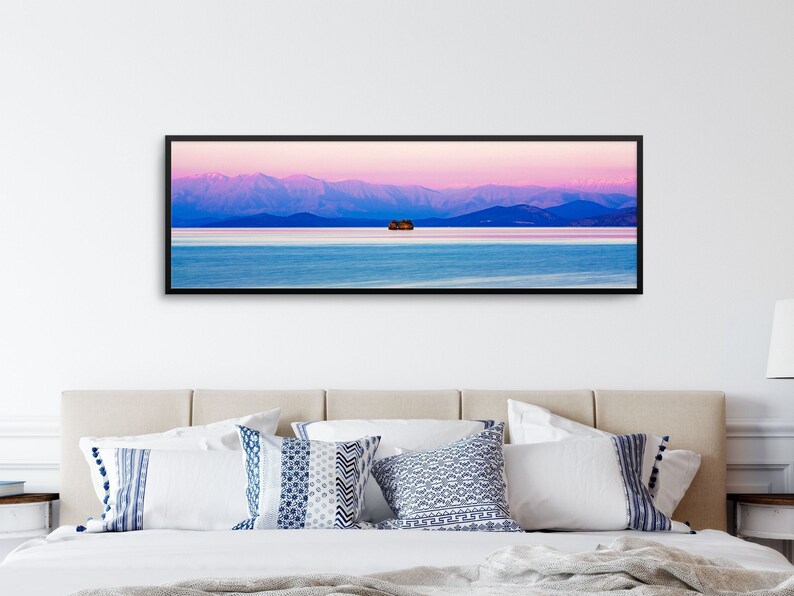 May include: A panoramic photograph of a mountain range in the distance, with a small island in the foreground. The sky is a vibrant pink and purple, reflecting in the calm water.