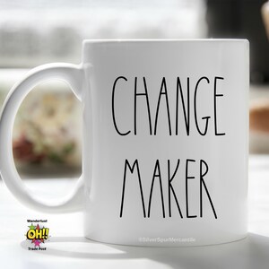 May include: White ceramic coffee mug with black text that reads "CHANGE MAKER". The mug has a handle and is sitting on a white surface.