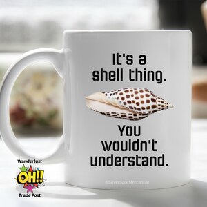 May include: White ceramic coffee mug with a black and brown seashell and the text "It's a shell thing. You wouldn't understand."