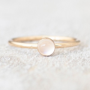 May include: A delicate gold ring featuring a round, white moonstone gemstone.