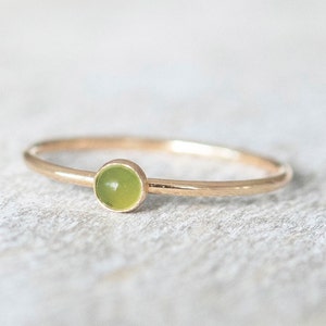 Puede incluir: Un delicado anillo de oro con una pequeña gema redonda de color verde claro. El anillo tiene una banda delgada y un diseño simple. La gema está engastada en un bisel de oro, creando una estética minimalista y refinada.