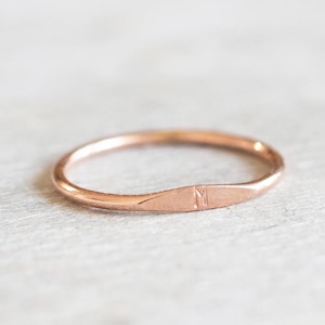 Peut inclure: Une simple bague en or rose avec un dessus plat légèrement incurvé. Le dessus de la bague est gravé de la lettre "M".