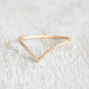 May include: A gold-colored V-shaped ring with a textured surface. The ring is set against a light-colored, textured background. The ring's design is simple and elegant, suitable for everyday wear.