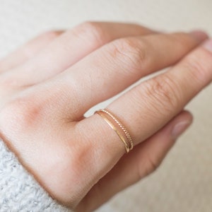 Op de afbeelding: Een close-up van een hand met twee dunne gouden ringen. De ring aan de onderkant is een eenvoudige band, terwijl de ring aan de bovenkant een gedraaide band is.