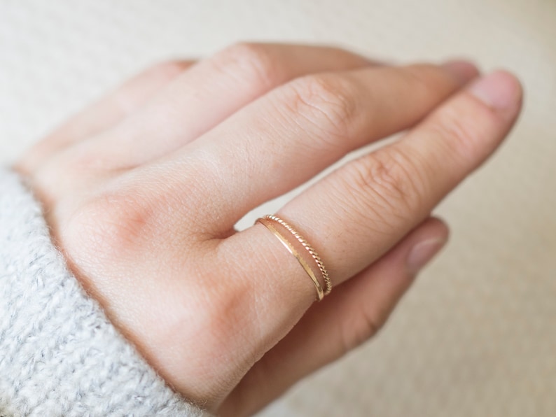 Op de afbeelding: Een close-up van een hand met twee dunne gouden ringen. De ring aan de onderkant is een eenvoudige band, terwijl de ring aan de bovenkant een gedraaide band is.