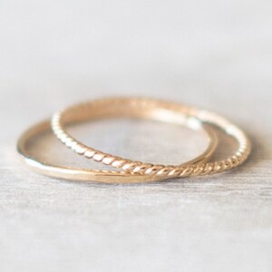 Op de afbeelding: Twee gouden ringen, een met een gladde afwerking en de andere met een gedraaid touwontwerp, liggen op elkaar.