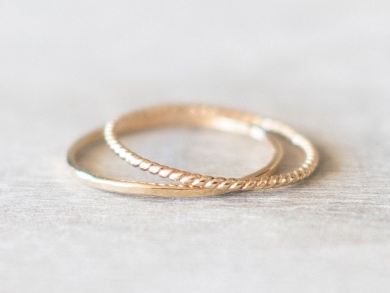 Op de afbeelding: Twee gouden ringen, een met een gladde afwerking en de andere met een gedraaid touwontwerp, liggen op elkaar.