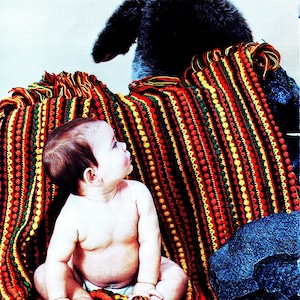 Könnte beinhalten: Ein Baby sitzt auf einer bunten gestreiften Häkeldecke mit einem Teddybären im Hintergrund. Die Decke hat rote, gelbe, grüne und schwarze Streifen mit einem strukturierten Muster.