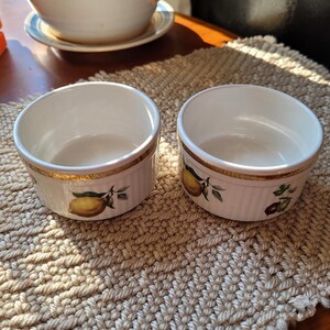 May include: Two white ceramic ramekins with a gold rim and fruit illustrations. The ramekins have a fluted design and are set on a woven placemat. The fruit illustrations include lemons and other fruits.