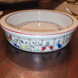 May include: White ceramic bowl with a red and blue rim. The bowl's exterior features a black and white design with illustrations of hand gestures and letters. The interior has a circular design with more letters and symbols.