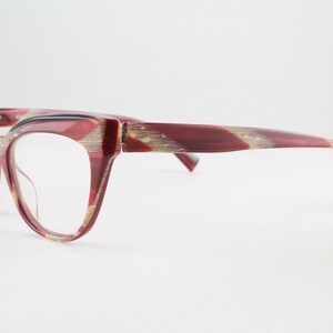 May include: Cat-eye glasses featuring a translucent deep red frame with subtle gold-toned horizontal streaks throughout the rims and temples. The glasses have clear lenses and a distinct upswept outer corner design on the frame front.