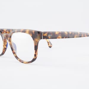 May include: Tortoiseshell eyeglasses featuring a thick, rounded frame with a prominent brow bar. The temples are connected to the frame front with a silver-toned hinge detail, and the clear lenses are set within the patterned acetate structure.
