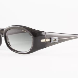 May include: Dark charcoal gray oval sunglasses with gradient lenses. The thick, glossy frames feature a silver-toned metal G logo emblem on each temple. The design includes a smooth, rounded bridge and wide arms that taper toward the ear tips.