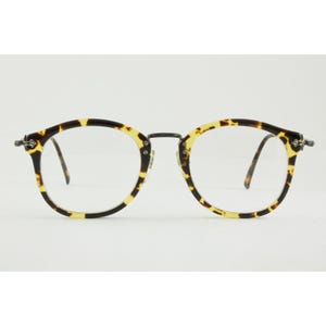 May include: Round eyeglasses featuring a tortoiseshell pattern frame with dark brown and yellow-gold mottled spots. The glasses have a thin dark metal bridge and matching metal temples, with small silver-toned rivets at the outer frame corners.