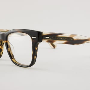 May include: Eyeglasses featuring a thick, dark brown and black tortoise shell pattern frame with clear lenses. The temples transition into a lighter, translucent beige and brown striped pattern, with small silver rectangular accents at the hinges.