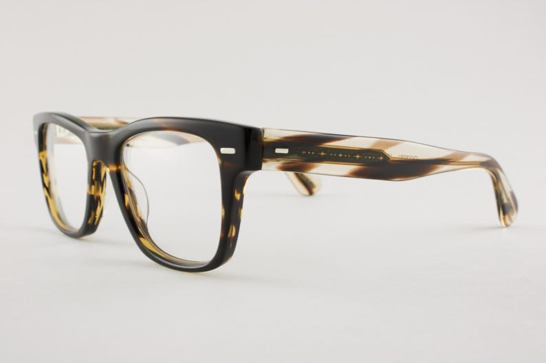 May include: Eyeglasses featuring a thick, dark brown and black tortoise shell pattern frame with clear lenses. The temples transition into a lighter, translucent beige and brown striped pattern, with small silver rectangular accents at the hinges.