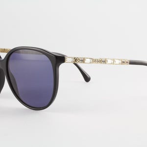 May include: Black sunglasses featuring large rounded frames with dark tinted lenses. The temples are gold-toned and embellished with a series of rectangular clear crystals along the sides, transitioning into black curved tips for a secure fit.