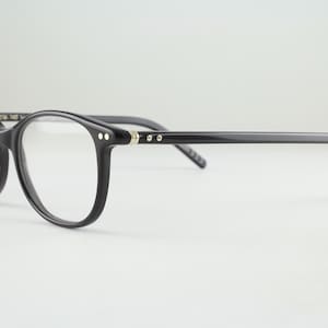 May include: A pair of black eyeglasses with a rounded frame shape. The frames feature a glossy finish and small silver-toned metal rivets on the front corners and hinges. The temples are thin and straight, extending from the frame front.