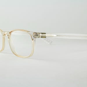 May include: Clear, transparent acetate eyeglasses with a rectangular frame shape. The glasses feature a subtle beige tint around the rims and clear temples. The hinges are metallic silver, and the frame has a smooth, polished finish throughout.