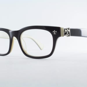 May include: Black and white eyeglasses featuring a rectangular frame design. The temples are accented with silver-toned metal fleur-de-lis emblems at the hinges. The front frame is black, while the inner side of the temples is white.