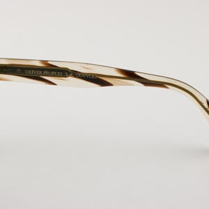 May include: Eyeglass frame temple arm featuring a tortoiseshell pattern with brown and cream tones. The inner side displays the text OLIVER PEOPLES and LA COVINA in gold lettering, with a metallic hinge connecting to the front frame section.