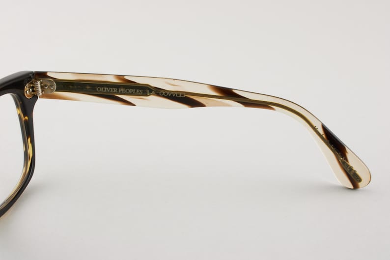 May include: Eyeglass frame temple arm featuring a tortoiseshell pattern with brown and cream tones. The inner side displays the text OLIVER PEOPLES and LA COVINA in gold lettering, with a metallic hinge connecting to the front frame section.