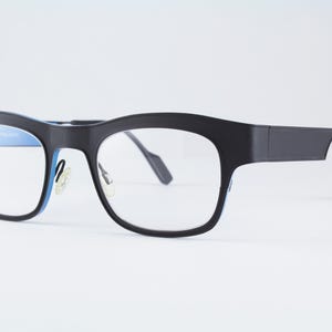 May include: Black eyeglasses with a thick frame and clear lenses. The inner rim of the frame features a bright blue accent color. The glasses have adjustable nose pads and thin, curved temples that extend from the sides of the frame.