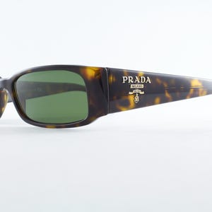 May include: Tortoiseshell-patterned sunglasses with green lenses. The Prada logo is displayed on the right temple. The sunglasses have a rectangular shape and are set against a white background.