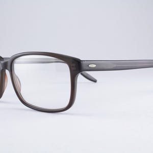 May include: Dark brown rectangular eyeglasses with clear lenses. The frames feature a smooth, matte finish with a small, silver-toned oval accent on the temple near the hinge. The bridge is curved, and the arms are straight with a slight downward curve.