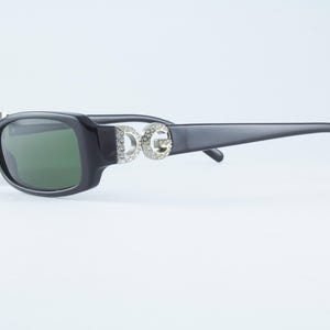 May include: Black sunglasses with green lenses. The frames are rectangular with a slight curve. The temples feature the letters "DG" embellished with rhinestones. The sunglasses are set against a white background.