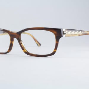 May include: Tortoiseshell-patterned eyeglasses with a rectangular frame shape. The temples feature a silver-toned metallic inlay with a repeating decorative pattern. The clear lenses are set within a brown and tan mottled acetate frame.