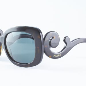 May include: Dark brown Prada sunglasses with a tortoiseshell pattern and a unique, curved temple design. The lenses are a muted blue-green color. The Prada logo is visible on the temple.