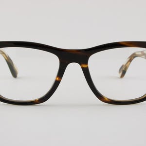 May include: Full-rim eyeglasses featuring a thick, dark brown and amber tortoiseshell pattern frame. The glasses have a rectangular shape with rounded edges, a keyhole bridge, and small silver-toned rectangular accents on the front corners.