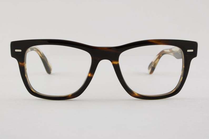 May include: Full-rim eyeglasses featuring a thick, dark brown and amber tortoiseshell pattern frame. The glasses have a rectangular shape with rounded edges, a keyhole bridge, and small silver-toned rectangular accents on the front corners.