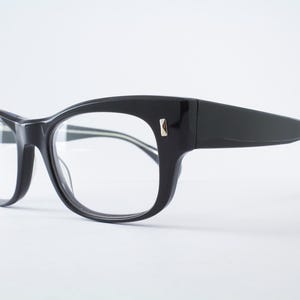 May include: Black eyeglasses with a thick, glossy frame and clear lenses. The frame features a distinct cat-eye shape with a small, metallic rectangular accent on the outer corners where the temples meet the front of the glasses.