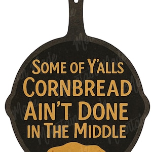 May include: A black, cast iron skillet-shaped sign with the text "SOME OF Y'ALLS CORNBREAD AIN'T DONE IN THE MIDDLE" in gold lettering. A golden cornbread shape is at the bottom. The sign has a handle.