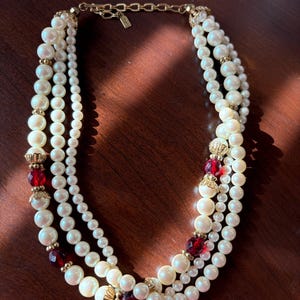 May include: A three-strand pearl necklace with gold accents and red jewel embellishments. The necklace features varying sizes of faux pearls and a gold chain clasp. The red jewels are clustered in decorative gold settings.