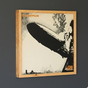 May include: Framed Led Zeppelin album art print. The artwork features a black and white image of a zeppelin with the band's name in orange at the top. The Atlantic Records logo is in the bottom right corner. The frame is made of light-colored wood.