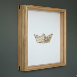 May include: A framed paper boat art piece. The boat is folded from a map, set against a white background. The frame is made of light-colored wood. The artwork is mounted on a dark gray wall.