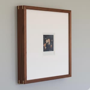 May include: A rectangular wooden picture frame with a white mat and a small photograph. The frame is made of dark brown wood with gold-colored accents at the corners. The photograph is a small, rectangular image of two people.