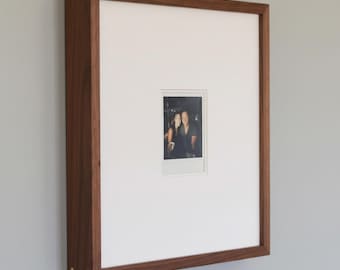 May include: A rectangular wooden picture frame with a white mat and a small photograph. The frame is made of dark brown wood with gold-colored accents at the corners. The photograph is a small, rectangular image of two people.
