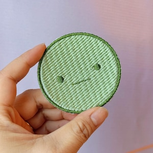 May include: A round, light green embroidered patch with a simple, slightly grumpy face. The patch has a textured appearance and a dark green border. The design features two small, dark eyes and a horizontal line for a mouth.